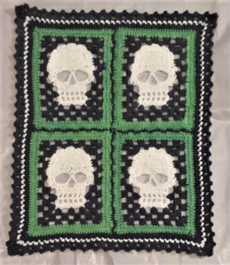 Puede incluir: Una manta de ganchillo con un esquema de color negro, verde y blanco. La manta presenta cuatro dise&ntilde;os de calaveras.