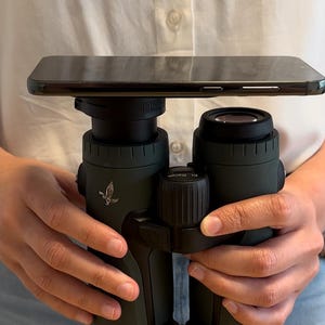 Op de afbeelding: Een donkergroene verrekijker met een smartphone erop. De verrekijker heeft een centrale focusknop en een logo. De smartphone is zwart en rechthoekig.