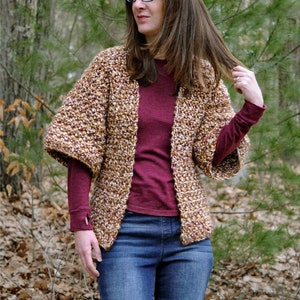 May include: A brown, white, and gold crocheted cardigan with long sleeves and a loose fit. The cardigan is open in the front and is being worn over a maroon long-sleeved shirt and blue jeans.