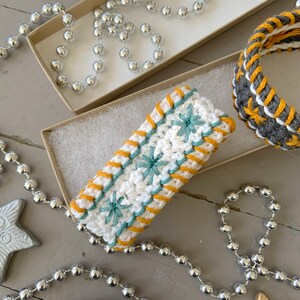 May include: A white crocheted bracelet with yellow and teal embroidery. The bracelet is in a box with white tissue paper and silver beads.