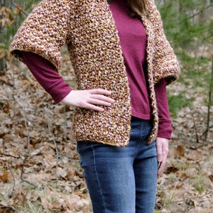 May include: A brown, white, and burgundy crocheted cardigan with a loose fit and 3/4 sleeves. The cardigan is worn over a burgundy long-sleeved shirt and blue jeans.