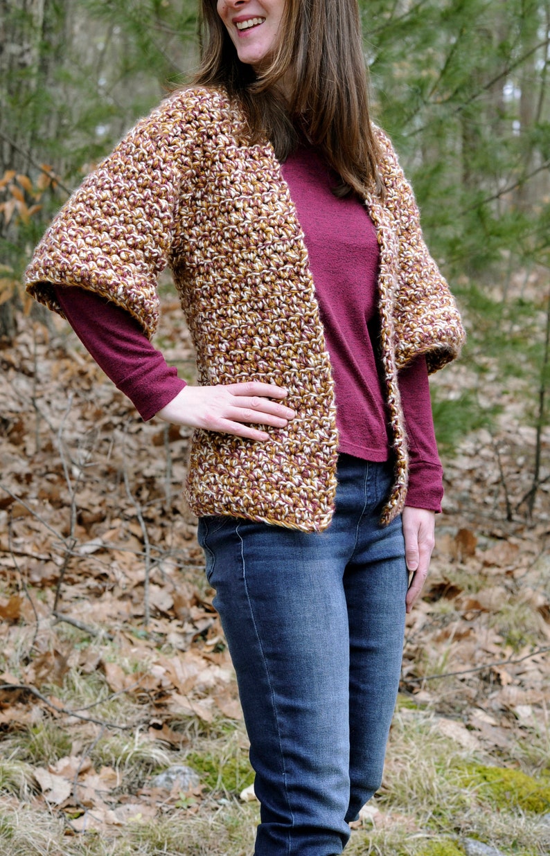 May include: A brown, white, and burgundy crocheted cardigan with a loose fit and 3/4 sleeves. The cardigan is worn over a burgundy long-sleeved shirt and blue jeans.