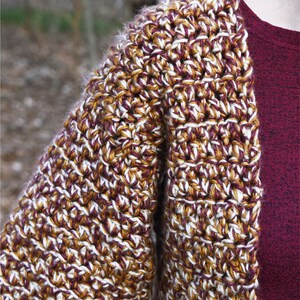 May include: A crocheted cardigan with a brown, white, and burgundy color pattern. The cardigan has a loose fit and a shawl collar.