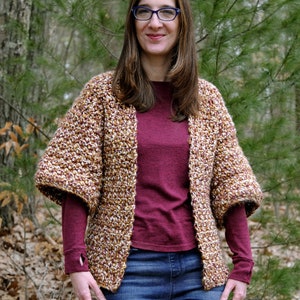 May include: A woman wearing a brown and white crocheted cardigan with a maroon long-sleeved shirt and blue jeans. She is standing in a wooded area.