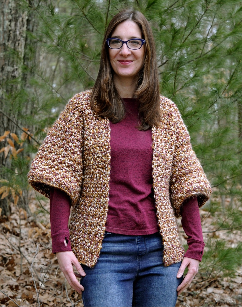 May include: A woman wearing a brown and white crocheted cardigan with a maroon long-sleeved shirt and blue jeans. She is standing in a wooded area.