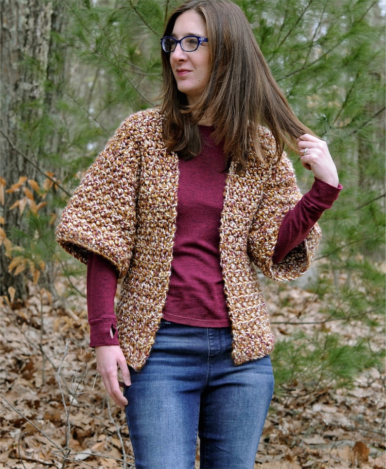 May include: A brown, white, and gold crocheted cardigan with long sleeves and a loose fit. The cardigan is open in the front and is being worn over a maroon long-sleeved shirt and blue jeans.