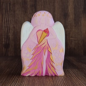 Pode incluir: Uma figura de anjo de madeira rosa com asas brancas, segurando um coração vermelho e dourado em suas mãos. O anjo está vestindo um vestido rosa com detalhes dourados e tem um design floral em suas asas.