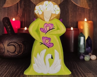 May include: A wooden figurine of a woman with a green dress, white flowers in her hair, and two purple butterflies on her dress. The figurine is standing on a wooden base and has a white swan carved on the front.