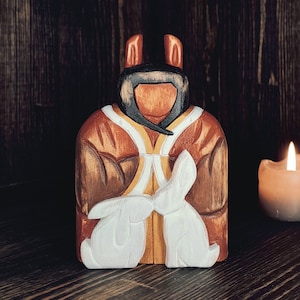 Puede incluir: Una figurita de madera de una persona que lleva una túnica marrón y blanca, sosteniendo dos conejos blancos en sus brazos. La figurita está de pie sobre una superficie de madera oscura.
