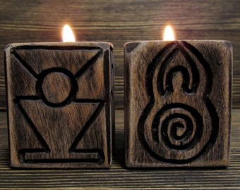 May include: Two square, dark brown wooden candle holders with carved symbols on the front faces. Each holder contains a lit candle with a visible flame. The left holder features a geometric design, while the right holder displays a spiral pattern.