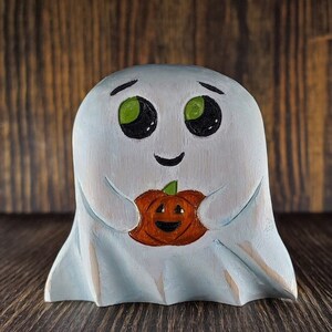 Pode incluir: Estatueta de fantasma de madeira azul claro segurando uma abóbora laranja com um rosto esculpido. O fantasma tem olhos verdes grandes e uma expressão sorridente. Decoração de Halloween.