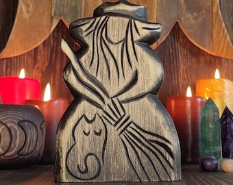 May include: A wooden carving of a witch holding a broom and a cat. The carving is dark brown and has a rustic look. The witch is wearing a pointed hat and has a long, flowing robe. The carving is set against a background of wooden planks and a triple moon symbol.