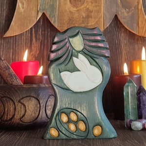 Könnte beinhalten: Eine Holzschnitzerei eines stilisierten Baumes mit einem weißen Kaninchen darin. Der Baum ist in Grün- und Brauntönen bemalt, mit einem lila und rosa Design an der Spitze. Die Schnitzerei steht auf einer Holzoberfläche mit Kerzen und Kristallen drumherum.