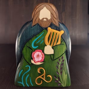 Pode incluir: Uma figura de madeira de uma pessoa com cabelo comprido e barba castanhos, vestindo uma túnica verde com guarnição azul e segurando uma harpa dourada. A túnica é decorada com uma flor, um símbolo triquetra e linhas azuis em espiral.