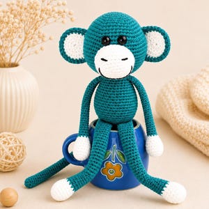 May include: A teal crochet monkey with white accents, sitting in a blue mug. The monkey has a white face, ears, and feet. The mug has a floral design. The monkey's arms and legs are long and slender.