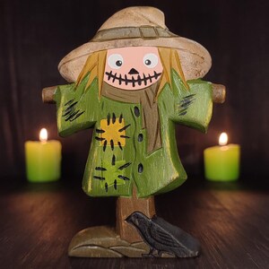 Pode incluir: Uma estatueta de espantalho de madeira com um rosto pintado, cabelo semelhante a palha e um casaco verde com remendos amarelos. O espantalho usa um chapéu castanho e tem uma boca costurada. Um corvo preto está na base. Duas velas verdes estão acesas no fundo.