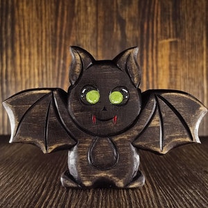 Pode incluir: Uma figura de morcego de madeira com olhos verdes e presas vermelhas. O morcego tem asas grandes e um rosto detalhado.