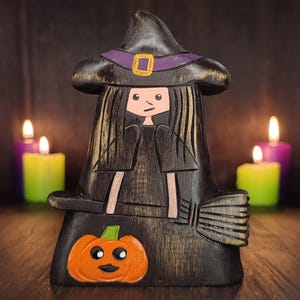 Pode incluir: Decoração de Halloween em madeira de uma bruxa com chapéu preto com faixa roxa e fivela dourada. A bruxa tem cabelo comprido preto e segura uma vassoura. Uma abóbora laranja com um rosto sorridente é esculpida na base.