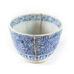 May include: A white and blue ceramic bowl with a geometric pattern. The bowl has been repaired with gold-colored kintsugi, a Japanese art form. The bowl is set against a white background.