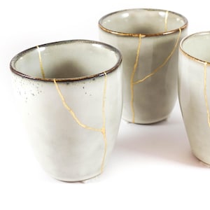 Może przedstawiać: Trzy kremowe ceramiczne kubki ze złotymi liniami naprawczymi kintsugi. Kubki mają lekko nakrapiany wygląd i ciemnobrązową obwódkę. Kubki są ułożone na białym tle.