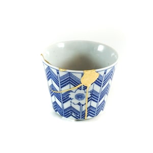 May include: A blue and white ceramic teacup with a gold crackle design. The cup has a geometric pattern and a small flower design in the center.