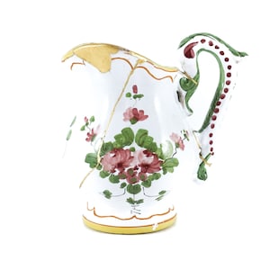 May include: A white ceramic pitcher with a floral design in shades of pink, green, and gold. The pitcher has a chipped handle and a crack on the side.