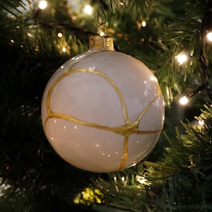 May include: White Christmas ornament with gold painted lines. The ornament is hanging on a Christmas tree.
