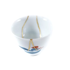 Op de afbeelding: Een witte theekop van keramiek met een blauw en rood bloemenmotief. De kop is gerepareerd met gouden kintsugi, een traditionele Japanse kunstvorm.
