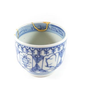 May include: A small, white ceramic teacup with blue decorative patterns and gold accents. The cup has a traditional design with a repaired crack, showcasing the Japanese art of Kintsugi.