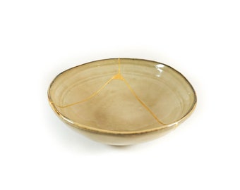 May include: A small, beige ceramic bowl with a crack that has been filled with gold paint.
