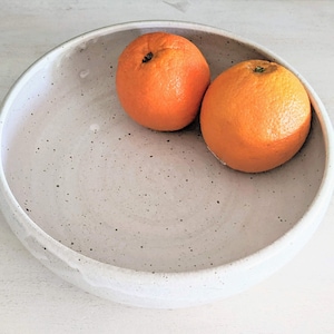 May include: A white ceramic bowl with a speckled glaze, featuring two whole oranges inside. The bowl has a slightly rounded rim and a smooth, textured surface.