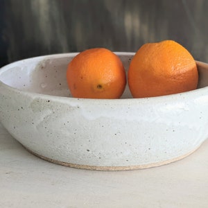 May include: A white ceramic bowl with a speckled glaze, filled with two bright orange oranges.