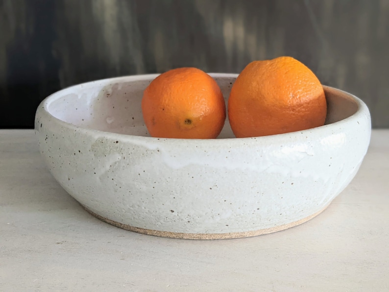 May include: A white ceramic bowl with a speckled glaze, filled with two bright orange oranges.