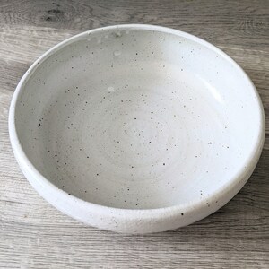 May include: A white ceramic bowl with a speckled glaze. The bowl is round and has a wide rim.