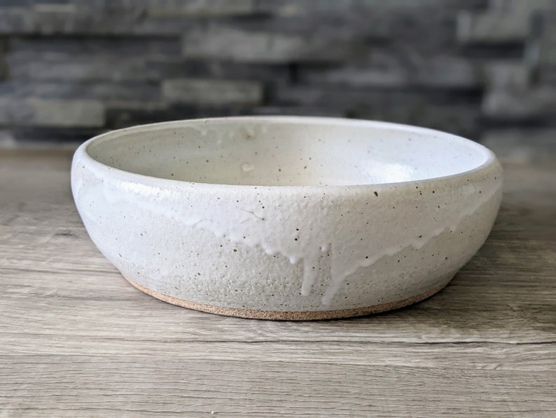 May include: A white ceramic bowl with a speckled glaze. The bowl has a rounded shape and a slightly uneven rim.