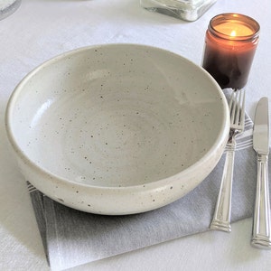 May include: A white ceramic bowl with a speckled glaze sits on a gray napkin with a silver fork and knife. A brown candle jar is in the background.