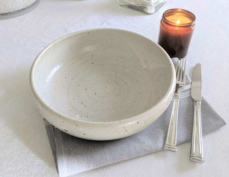 May include: A white ceramic bowl with a speckled glaze sits on a gray napkin with a silver fork and knife. A brown candle jar is in the background.