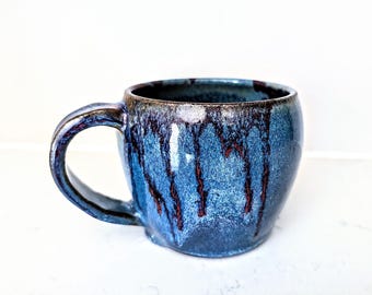 May include: A handmade ceramic mug with a rounded body and a curved handle. The mug features a glossy, drippy glaze in shades of blue with streaks of reddish-brown. The interior is a darker blue, and the mug sits on a white surface.