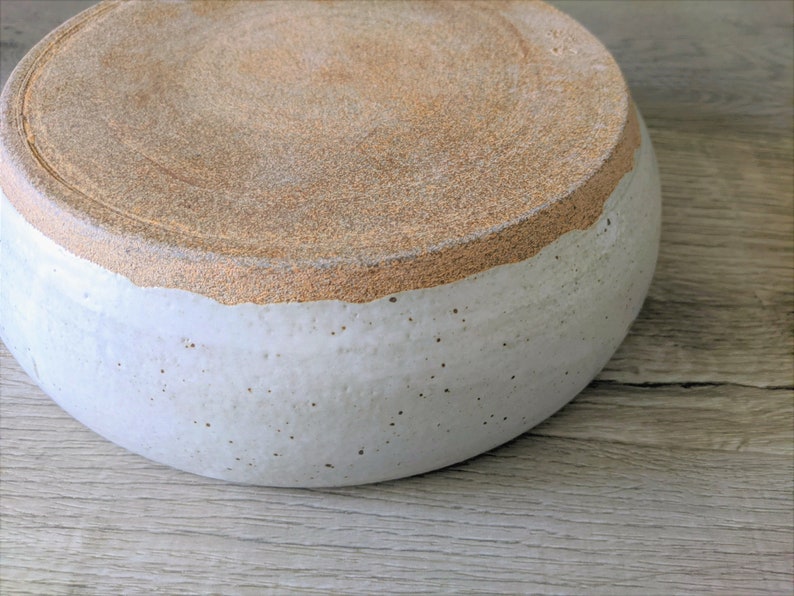May include: A large, white ceramic bowl with a brown rim. The bowl is slightly speckled and has a rough texture.