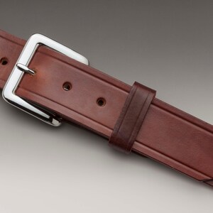 May include: A brown leather belt with a silver buckle. The belt has a stitched design and multiple holes for adjusting the fit.