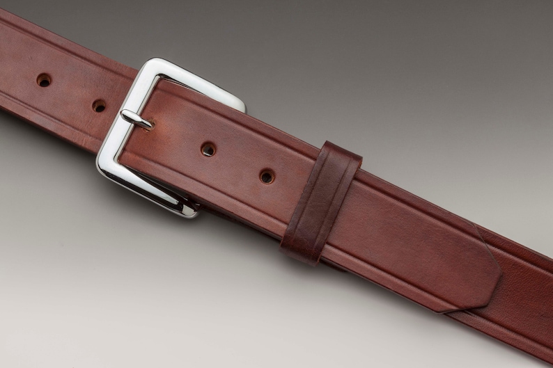 May include: A brown leather belt with a silver buckle. The belt has a stitched design and multiple holes for adjusting the fit.