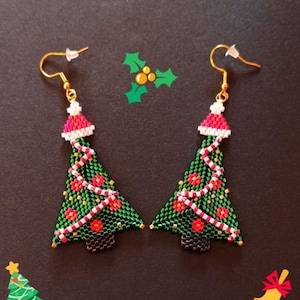 Peut inclure: Une paire de boucles d'oreilles en forme de sapin de Noël, faites de perles vertes, rouges et blanches. Chaque boucle d'oreille présente un bonnet de Père Noël rouge et blanc, une guirlande de perles et une base marron. Le texte "Aux Perles Enchantées" est visible.