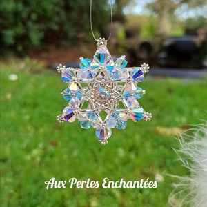 Puede incluir: Un adorno de copo de nieve plateado con cristales azules iridiscentes y transparentes. El adorno está colgado de una cuerda plateada y se encuentra frente a un fondo de césped verde. El texto "Aux Perles Enchantées" es visible en la parte inferior de la imagen.