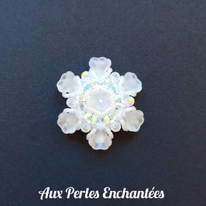 Peut inclure: Une décoration en forme de flocon de neige en perles blanches avec des perles irisées et une perle centrale transparente. La décoration est sur un fond bleu foncé.