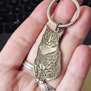 May include: A silver-toned metal keyring featuring a laser-engraved portrait of a tabby cat. The cat is depicted in a seated position with detailed fur markings and visible whiskers. The charm is attached to a circular split ring made of polished metal.