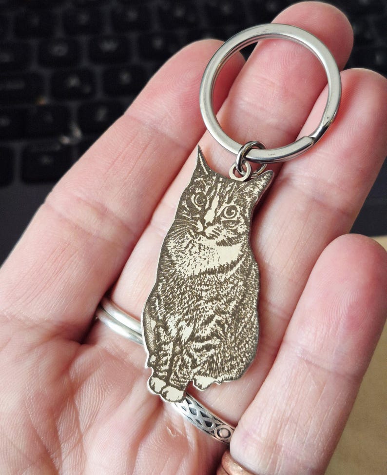 May include: A silver-toned metal keyring featuring a laser-engraved portrait of a tabby cat. The cat is depicted in a seated position with detailed fur markings and visible whiskers. The charm is attached to a circular split ring made of polished metal.