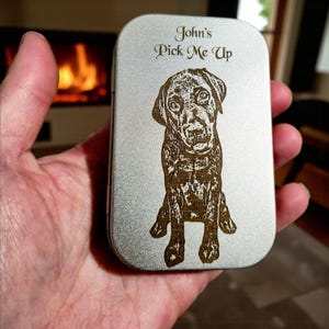 Puede incluir: Una caja metálica plateada con una imagen grabada detallada de un perro Labrador Retriever. El texto "John's Pick Me Up" está encima del perro. La caja está sujeta en la mano de una persona.
