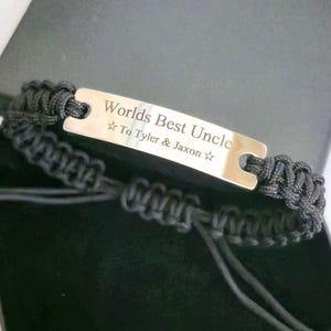 Könnte beinhalten: Ein schwarzes Makramee-Armband mit einer silbernen rechteckigen Platte. Auf der Platte stehen die Worte "Worlds Best Uncle" und die Namen "Tyler & Jaxon". Das Armband wird in einer schwarzen Box präsentiert.