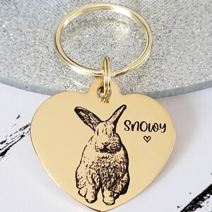 May include: Gold heart-shaped pet ID tag with a black and white engraved image of a rabbit and the name "Snowy" with a small heart symbol.