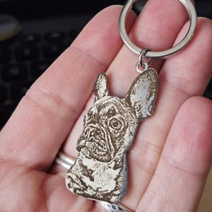 May include: A silver-toned metal keyring featuring a laser-engraved portrait of a French Bulldog. The charm is shaped to the outline of the dog's head and ears, attached to a circular split ring. The engraving displays dark, detailed shading.